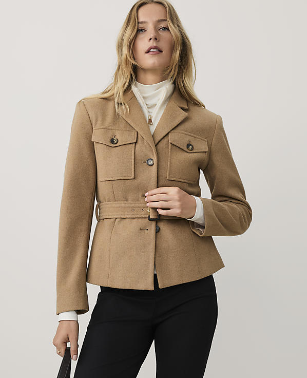 A woman wearing a tan belted jacket, white turtleneck, and black trousers stands against a plain light gray background.
