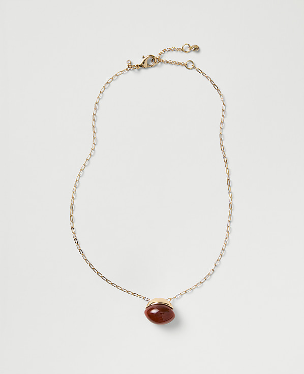 A gold chain necklace with a red oval gemstone pendant rests against a plain light gray background.