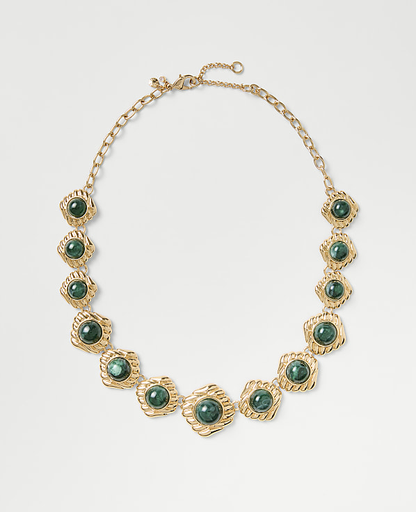 A gold chain necklace featuring green circular gemstones is centered against a plain light gray background.