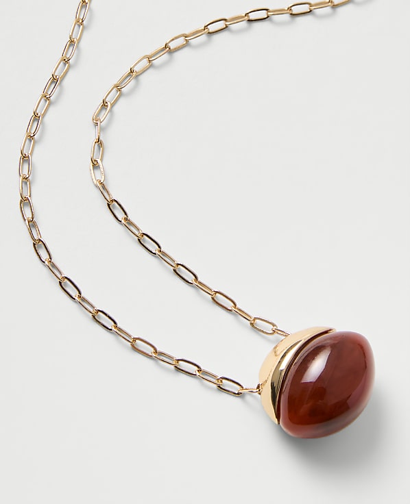 A gold chain necklace with a large, oval red gemstone pendant rests on a plain white background.