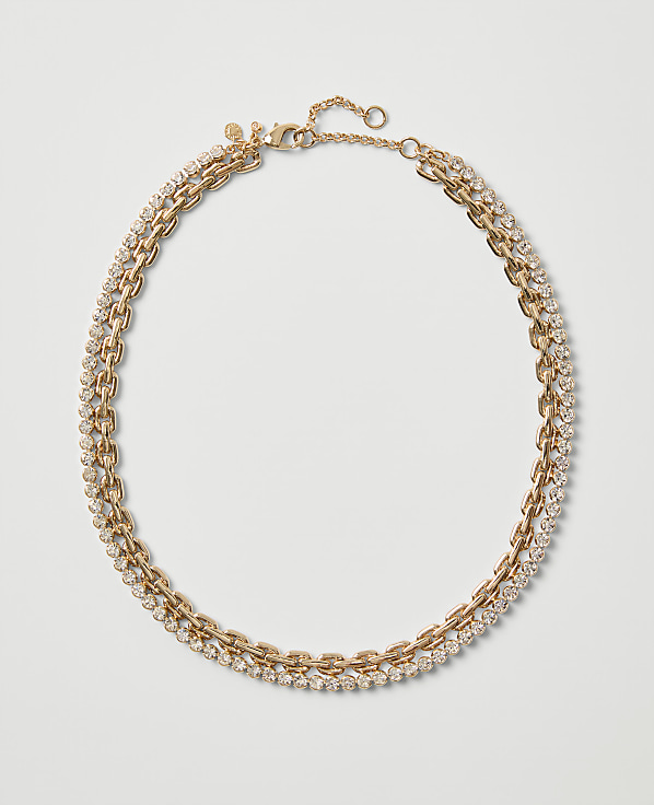A gold-toned chain necklace embellished with sparkling crystals rests on a plain, light gray background.