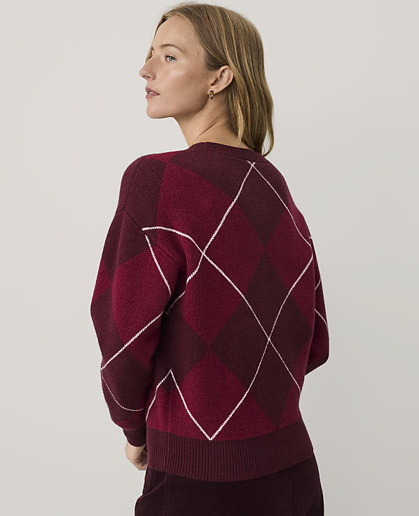 A woman with blonde hair wearing a burgundy argyle sweater stands against a plain light gray background.