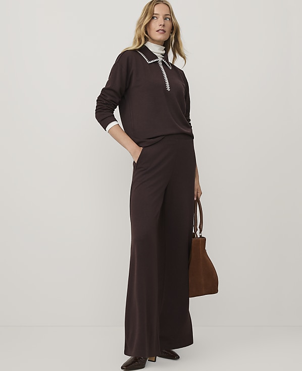 A woman wearing a dark brown two-piece outfit and holding a brown handbag stands against a plain white background.