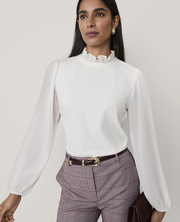 A woman wearing a white ruffled blouse and plaid trousers stands against a plain, light-colored background.
