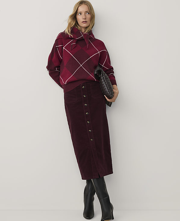 A woman wearing a red plaid sweater and a long burgundy skirt stands against a plain white background.