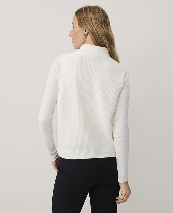 A woman wearing a white ribbed sweater and black pants stands against a plain, light-colored background.