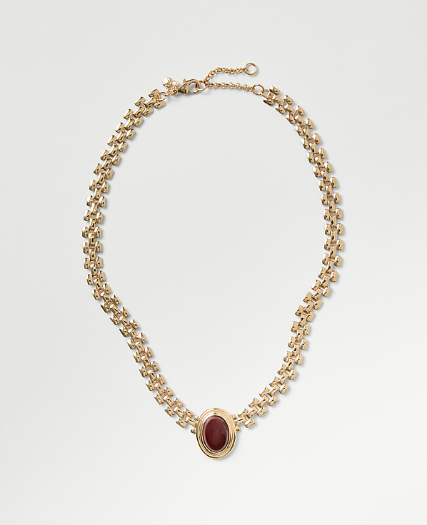A gold chain necklace with a red oval gemstone pendant rests on a plain light gray background.