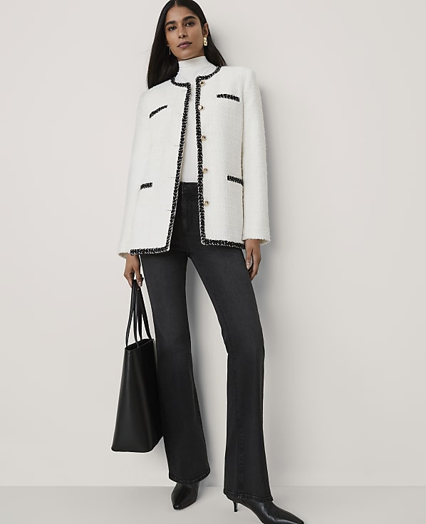 A woman wearing a white tweed jacket, black flared jeans, and holding a black tote bag stands against a plain light gray background.