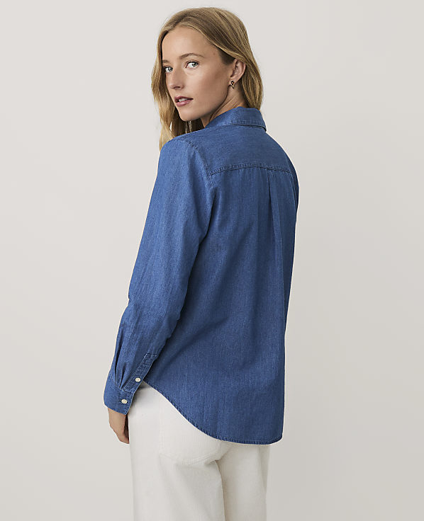 A blonde woman wearing a blue denim shirt and white pants stands against a plain, light-colored background.