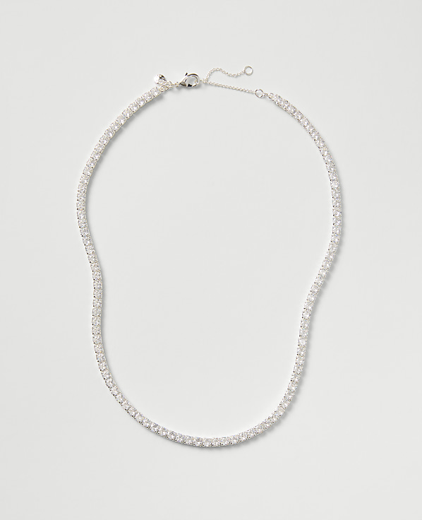 A sparkling tennis necklace with clear stones rests against a plain, light gray background.
