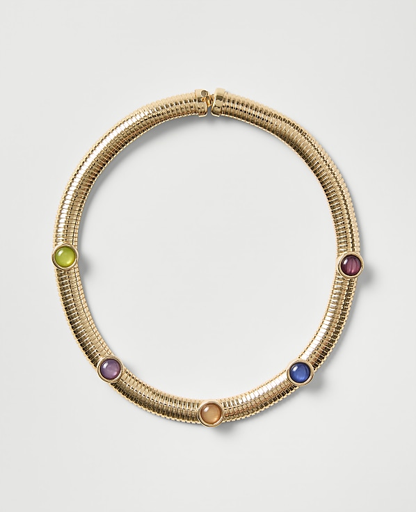 A gold ribbed necklace featuring five colorful gemstones is centered against a plain light gray background.