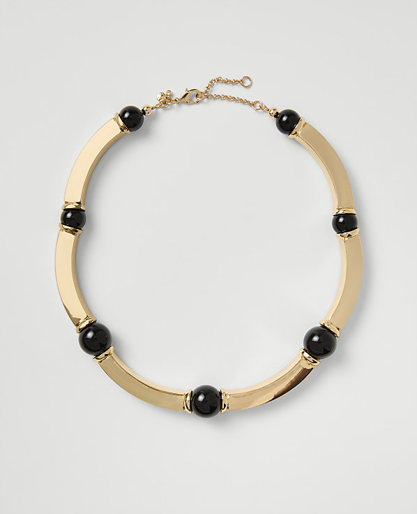 A gold-toned necklace with black spherical beads rests on a plain light gray background.