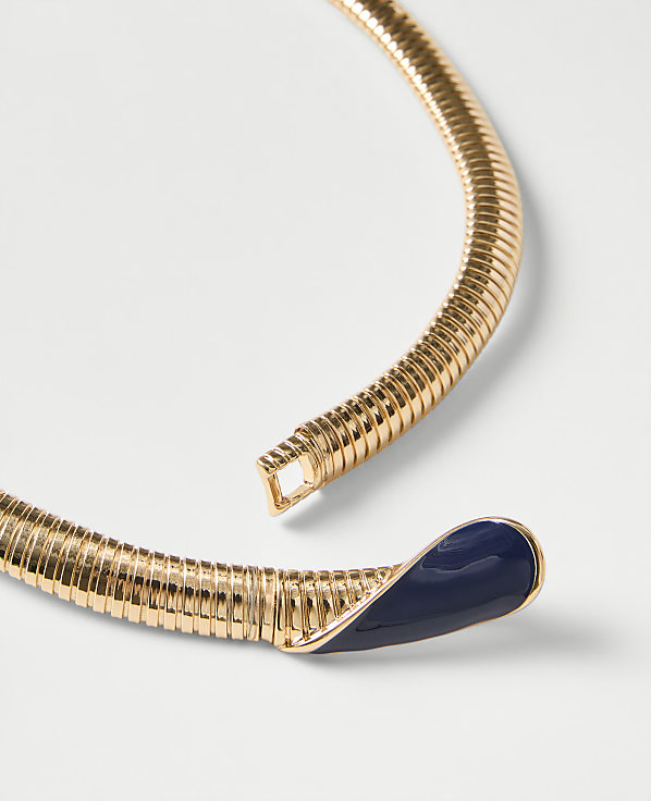 A gold-toned ribbed collar necklace with a navy blue enamel tip rests on a plain white background.