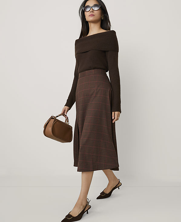 A woman wearing a brown off-the-shoulder top, a plaid skirt, and sunglasses stands against a plain white background.