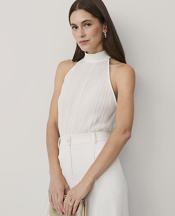 A woman wearing a white pleated halter top and white trousers stands against a plain, light-colored background.