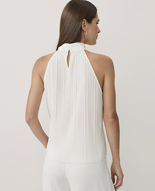 A woman wearing a white pleated halter top and white trousers stands against a plain light gray background.