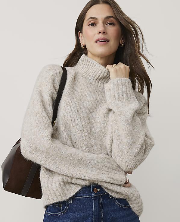 A woman wearing a beige turtleneck sweater and blue jeans stands against a plain, light-colored background.