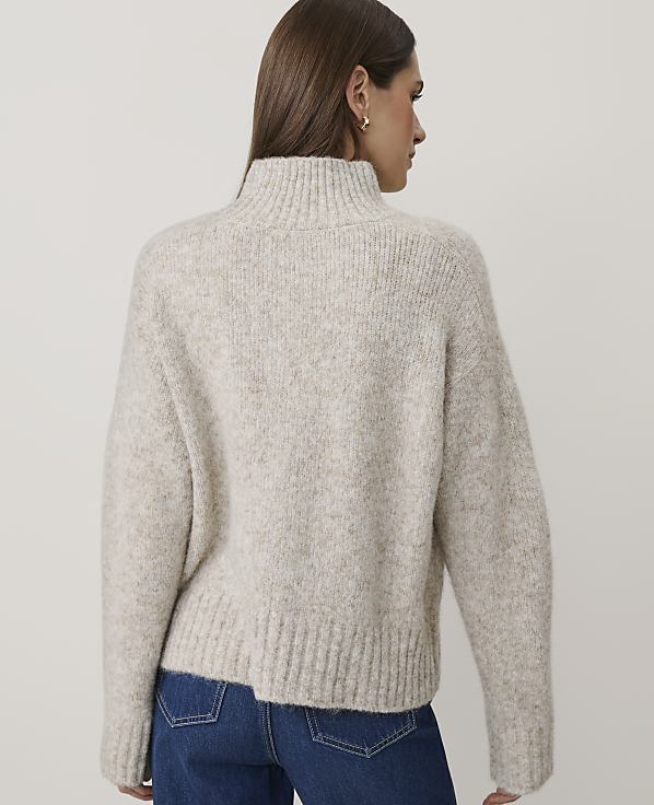 A woman wearing a beige knit sweater and blue jeans stands against a plain, light-colored background.