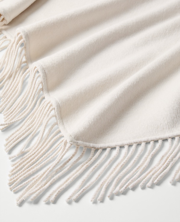 A cream-colored fringed blanket is draped in soft folds over a plain white background.
