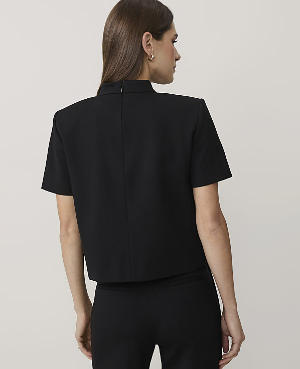 A woman wearing a black short-sleeved top and matching trousers stands with her back to the camera against a plain, light-colored background.