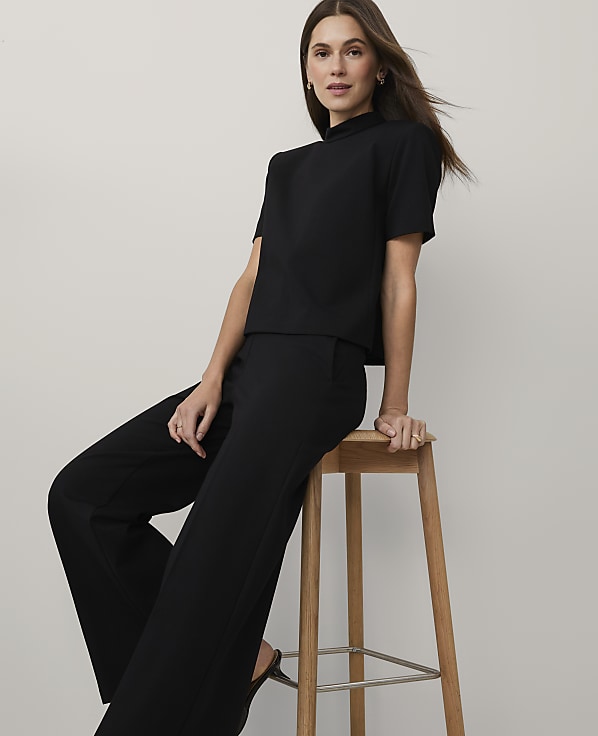 A woman wearing a black short-sleeved top and wide-leg trousers sits on a wooden stool against a plain light gray background.