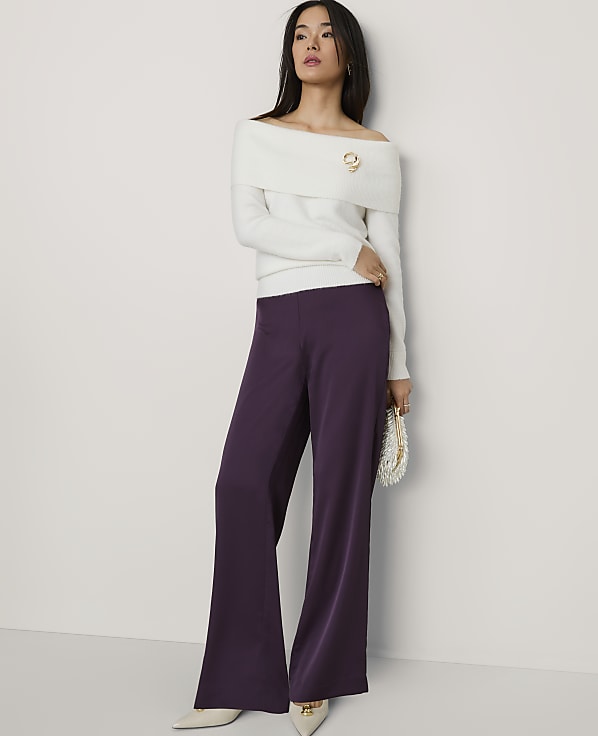 A woman wearing a white off-the-shoulder sweater and wide-leg purple trousers stands against a plain white wall.