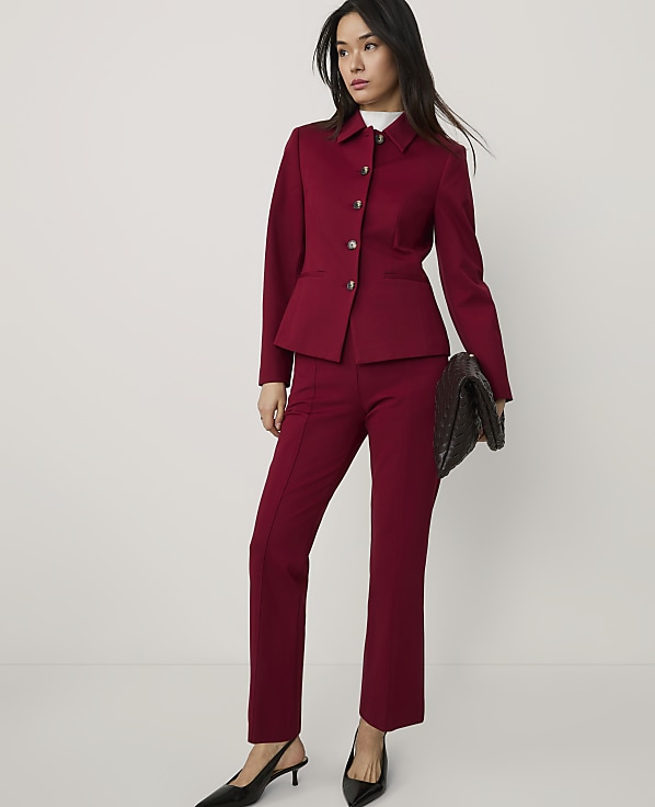 A woman wearing a burgundy suit and black heels stands against a plain white background.