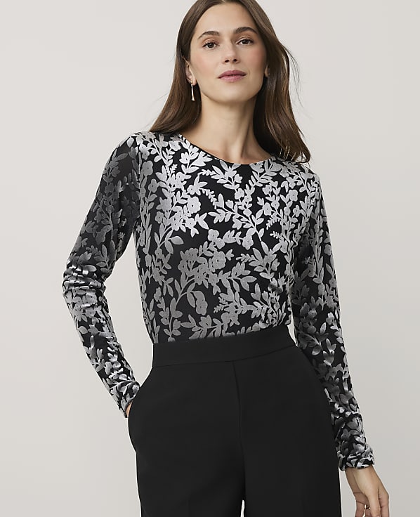 A woman wearing a silver floral print long-sleeved top and black trousers stands against a plain light gray background.