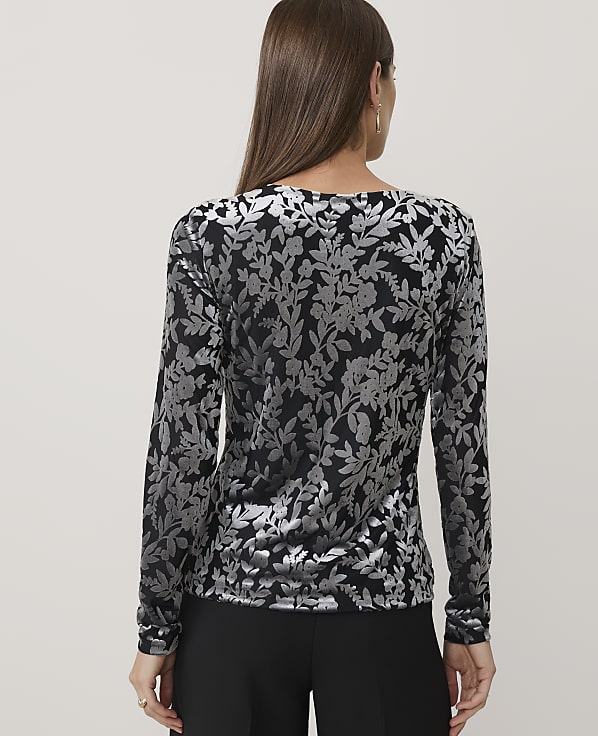 A woman wearing a black and silver floral print top and black trousers stands against a plain light grey background.