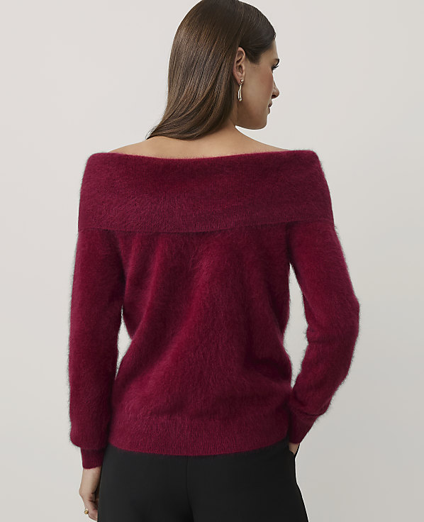 A woman wearing a fuzzy burgundy off-the-shoulder sweater and black pants stands with her back to the camera against a plain light gray background.