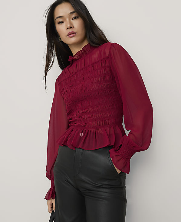 A woman wearing a smocked red blouse and black leather pants stands against a plain light gray background.