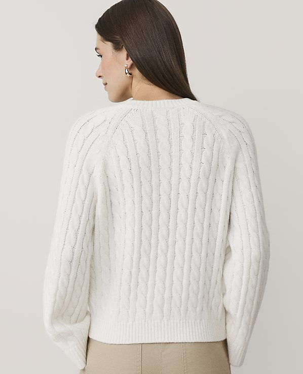 A woman wearing a white cable-knit sweater and beige pants stands with her back to the camera against a plain, light-colored background.