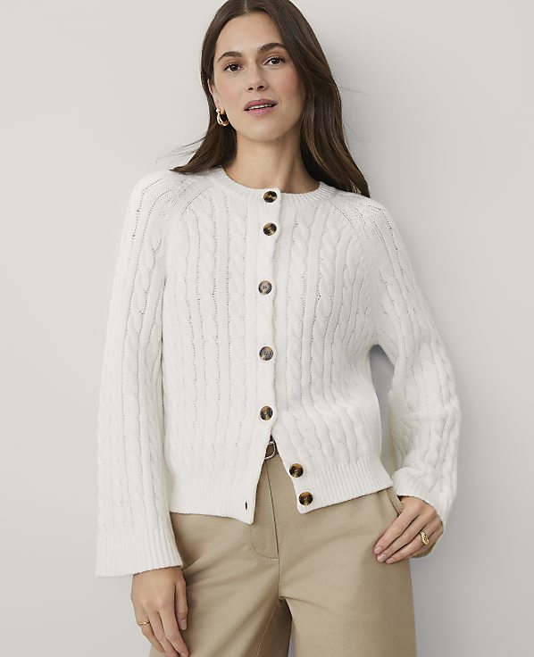 A woman wearing a white cable-knit cardigan and khaki pants stands against a plain, light-colored wall.