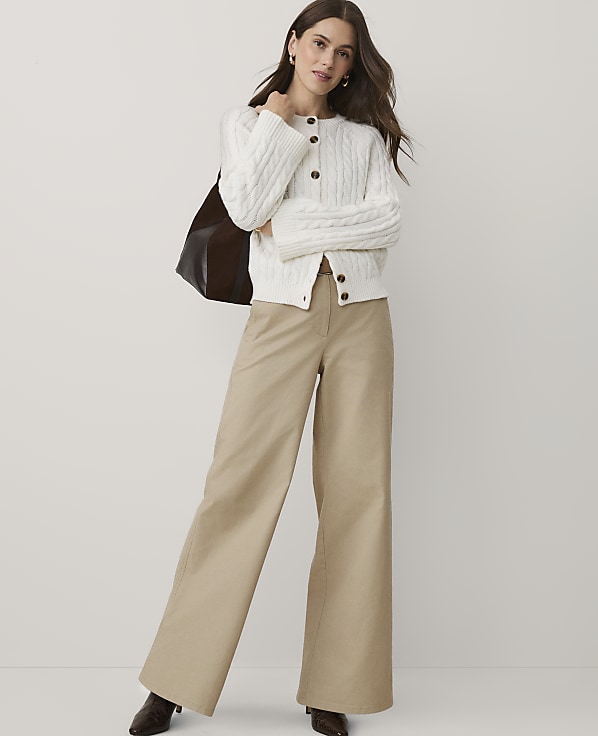 A woman wearing a white cable-knit cardigan and wide-leg khaki trousers stands against a plain white background.