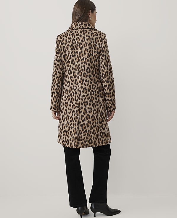 A woman wearing a leopard-print coat and black trousers stands with her back to the camera against a plain white background.