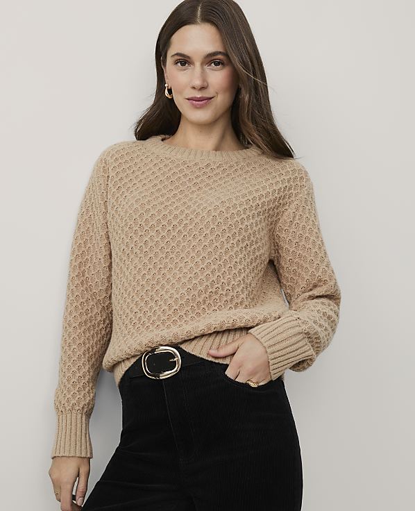 A woman wearing a tan textured sweater and black corduroy pants stands against a plain light gray background.