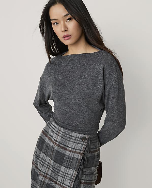 A woman wearing a grey long-sleeved top and a plaid skirt stands against a plain, light-colored background.