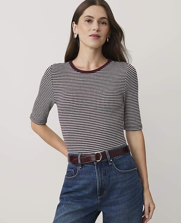 A woman wearing a purple and white patterned top and blue jeans stands against a plain, light-colored background.