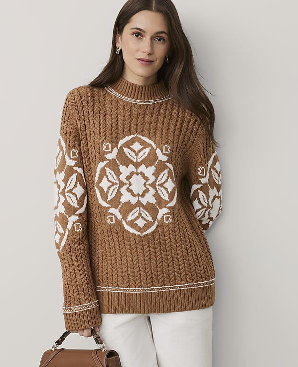 A woman wearing a brown cable-knit sweater with white floral patterns and white pants stands against a plain, light-colored background.