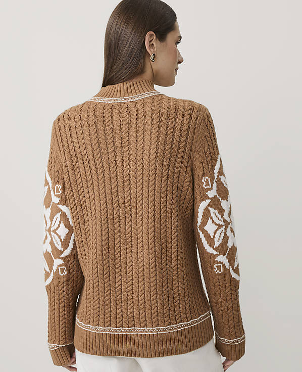 A woman stands with her back to the camera wearing a brown cable-knit sweater with white patterns on the sleeves against a plain white background.