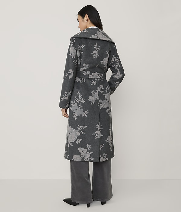 A woman stands with her back to the camera wearing a long, grey floral-patterned coat and wide-leg trousers against a plain, light-colored background.