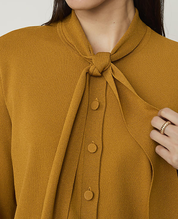 A person wears a mustard-yellow knit cardigan with a tie-neck detail against a plain white background.