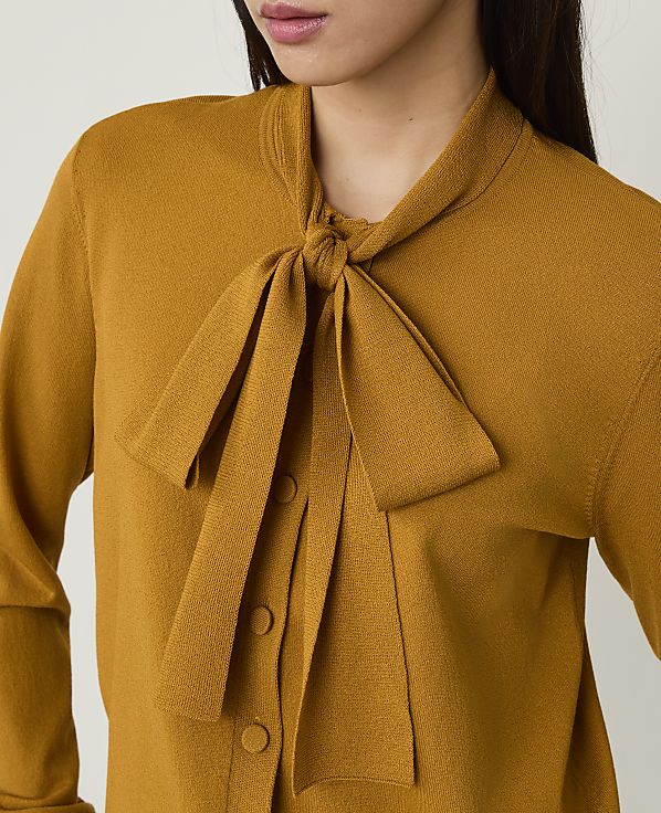 A person wears a mustard-yellow knit top with a large bow tie against a plain white background.