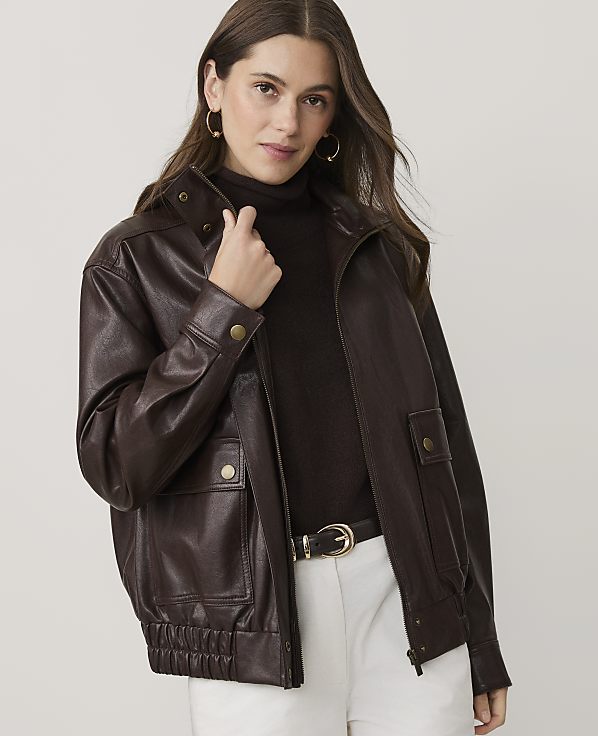 A woman wearing a dark brown leather jacket, a brown turtleneck, and white pants stands against a plain, light-colored background.
