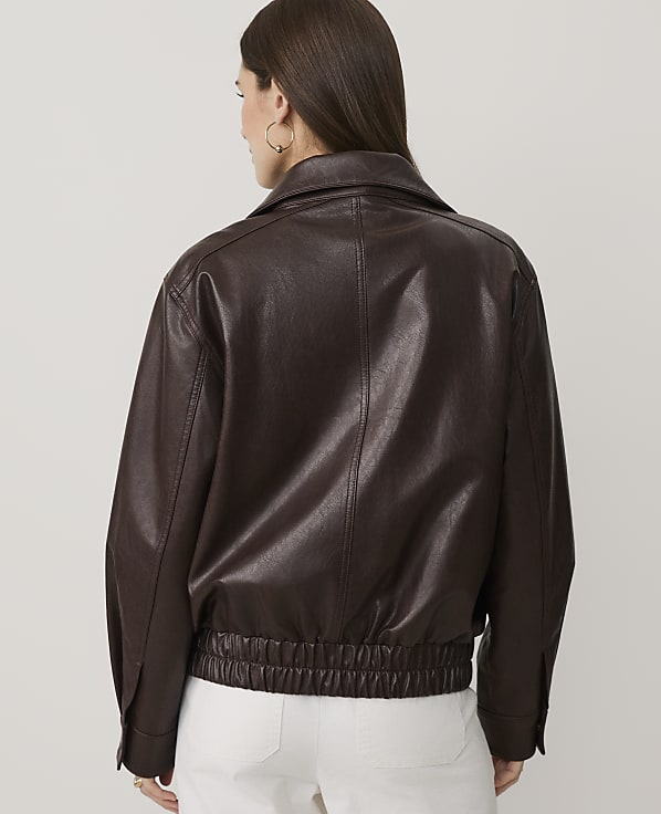 A woman wearing a dark brown faux leather jacket and white pants stands with her back to the camera against a plain light gray background.