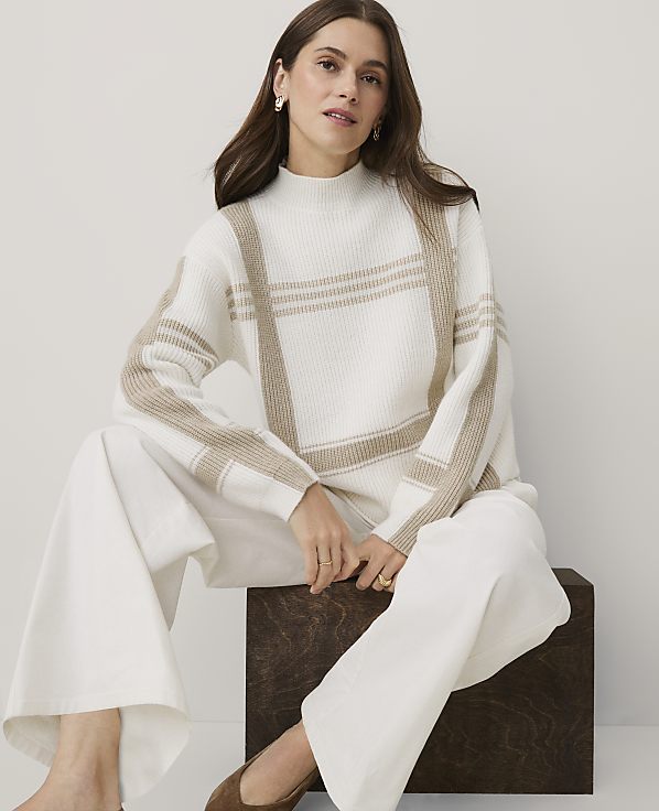 A woman wearing a white and beige patterned sweater and white trousers sits on a dark wooden block against a plain light grey background.