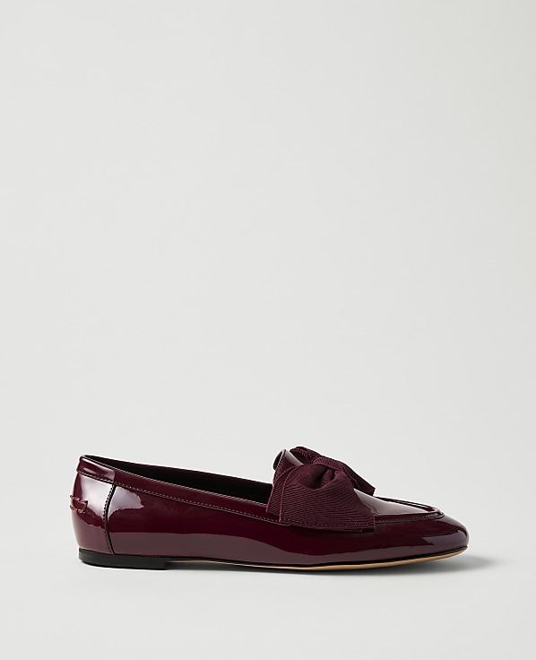 A burgundy patent leather flat shoe with a bow sits in the foreground against a plain, light gray background.