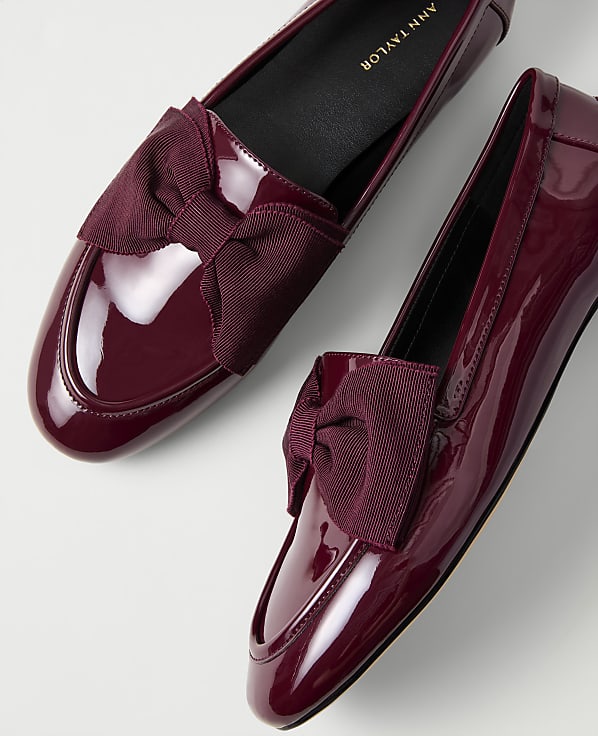 A pair of glossy burgundy patent leather loafers with bow accents are set against a plain white background.