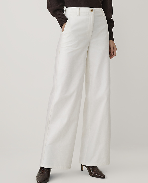 A person wearing a brown sweater, wide-leg white trousers, and dark brown heels stands against a plain, light-colored background.