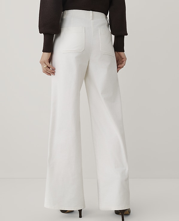 A person wearing a dark brown sweater and wide-leg white trousers stands against a plain light gray background.